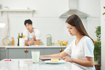Young Asian woman in a white T-shirt sitting at a kitchen dining table chair, reading while eating bread and drinking milk
