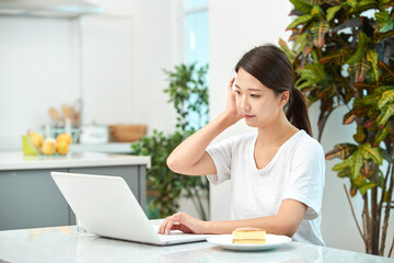 Fototapeta premium Young Asian woman in a white T-shirt sitting at a kitchen dining table chair, working on a laptop while eating bread and drinking milk
