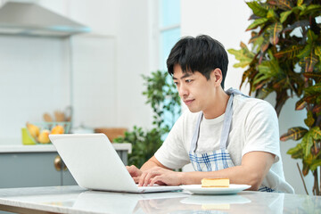 Young Asian man in a white T-shirt and apron sitting at a kitchen dining table chair, working on a laptop while eating bread