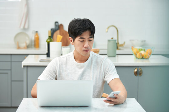 Young Asian man in a white T-shirt sitting at a kitchen dining table chair in a living room, working on a laptop and using a smartphone