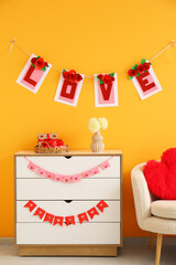 Chest of drawers with buntings, gift boxes and flowers near orange wall. Valentine's Day celebration