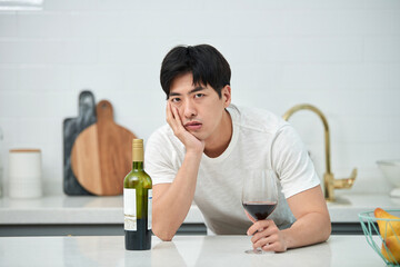 A young Asian man at the kitchen table holding a wine bottle and a wine glass, struggling with a...