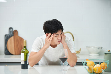 A young Asian man at the kitchen table holding a wine bottle and a wine glass, struggling with a...