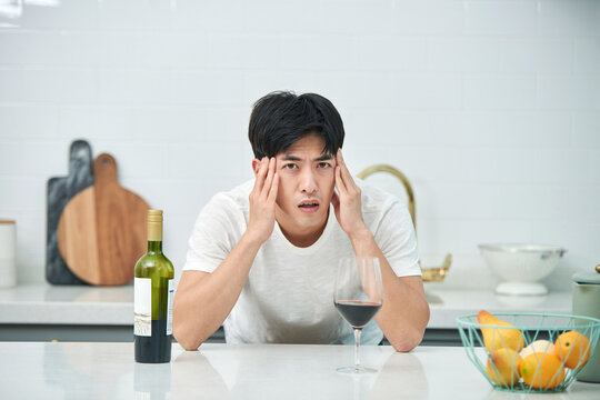 A young Asian man at the kitchen table holding a wine bottle and a wine glass, struggling with a hangover from the previous day.