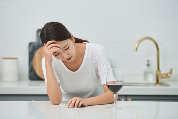 A young Asian woman at the kitchen table holding a wine glass, struggling with a hangover from the...