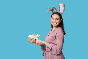 Happy young woman in bunny ears with sweet Easter cake and toy rabbit on blue background