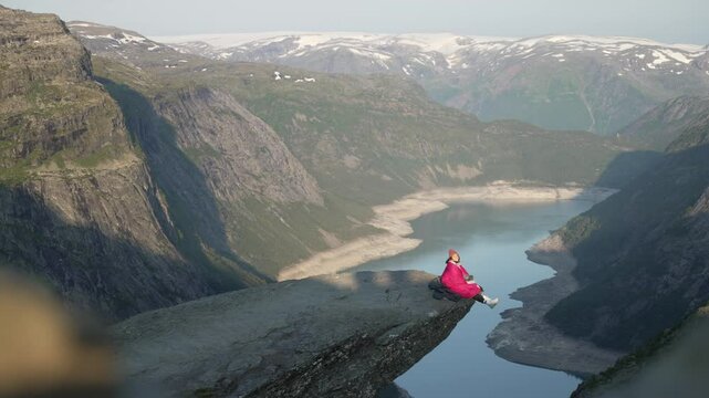 Girl wrapped in sleeping bag is sitting on the edge of cliff on Trolltunga, Norway. Aerial of Norwegian Troll Tongue.