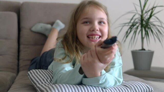 Caucasian child girl laying on sofa home and watching tv,using pilot,remote control.Happy kid indoors have a fun.Lesure concept.