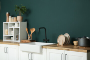 Blurred view of modern kitchen with white counters, sink and utensils near green wall