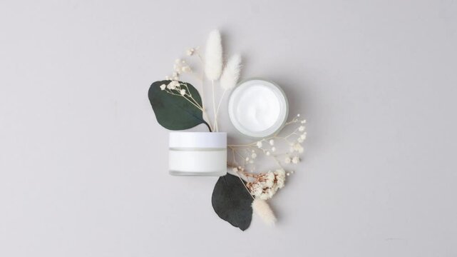 Jars of cosmetic cream with eucalyptus and flower on grey background. Flat lay, copy space