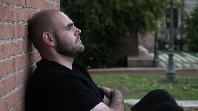 A young bald man sits on the ground, looking perplexed and sad, lost in deep thoughts. His posture reflects a sense of hopelessness.