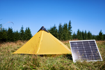 Yellow tent with solar panel set up in grassy field surrounded by dense evergreen trees under clear...