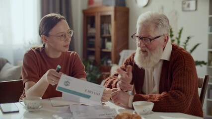 Young woman and her elderly grandfather sitting at table in living room, reviewing bill and discussing household expenses during day at home