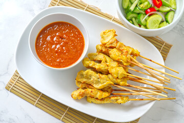 Moo Satay is a Thai-style grilled pork skewer with peanut sauce and cucumber salad. It is a top-view Thai food snack Served on a white plate on a bamboo mat with wooden skewers.