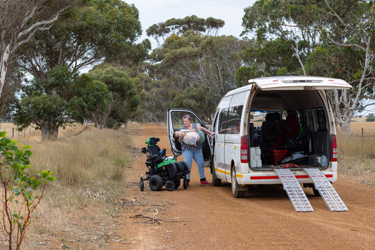 Mother transferring adult son from van to wheelchair on a gravel access road in country