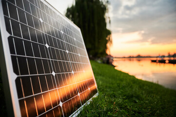 Solar panel illuminated by setting sun stands on grassy lakeside, with picturesque sunset and trees...