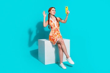 Full size photo of pretty young girl sit cube arm wave make selfie phone wear dress isolated on teal color background