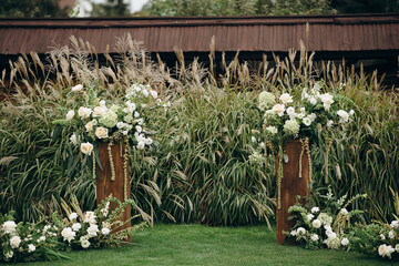 Two wooden pedestals with white floral arrangements stand amidst tall pampas grass in an outdoor...