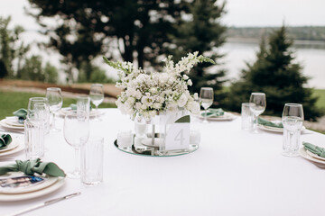 A white floral centerpiece in a modern vase on a white tablecloth in an outdoor setting