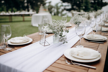 Elegant outdoor wedding table setting with white tablecloth, glassware, and gypsophila floral arrangements in clear vases. Wooden table and blurred greenery create a natural and romantic atmosphere