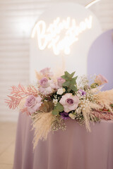 A delicate floral arrangement of lavender roses and pampas grass adorns a round table with a mauve tablecloth. A glowing neon sign in the blurred background