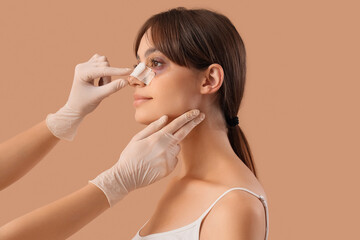 Surgeon hands touching woman's bandaged nose after plastic surgery on beige background