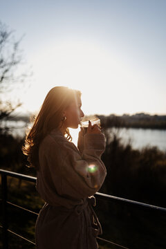 Full shot of a woman in a beige robe, drinking from a clear glass, outdoors on a balcony at sunset.