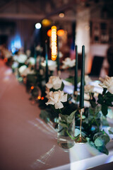 Many thin green candles in slender single candle holders surrounded by flowers on a banquet table in a restaurant, featuring white and green tones for a celebration or banquet