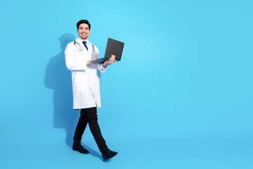 Confident Male Doctor Standing on a Blue Background Holding a Laptop and Smiling in a Medical Uniform
