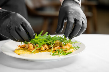 A chef in black gloves carefully garnishes a gourmet dish featuring an omelette topped with shrimp and fresh arugula. The elegant presentation highlights culinary art.