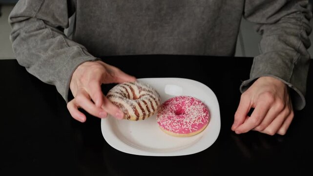 Close-up of two donuts on a plate as hands hesitate, touch, and examine them, struggling to decide which one to eat. Pink frosted donut with white sprinkles