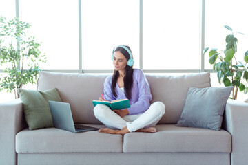 Fototapeta premium Young woman sitting comfortably on a sofa wearing headphones while writing in a notebook and using a laptop