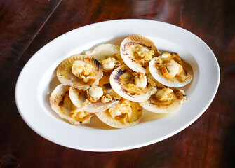 Grilled scallops in the shell with butter, a fresh seafood menu of a restaurant, served on a white plate on the wooden table with Thai seafood sauce.