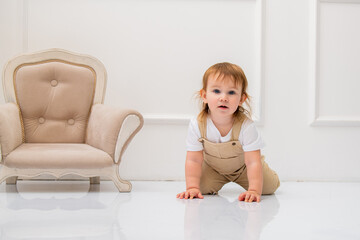 young toddler in beige overalls and a white t-shirt crawls on a glossy white floor, gazing curiously at the camera. A stylish vintage armchair sits beside the child, complementing the elegant minimali