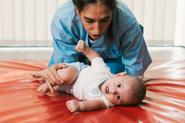 Physiotherapist assisting baby with assisted rolling exercises on red mat