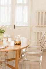 Fototapeta premium A cozy and inviting dining environment showing a round wooden table set with tea cups, tea pot, and modern transparent chairs, with soft natural lighting and flowers adding charm.