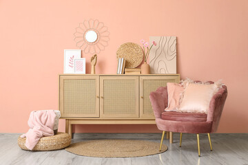 Interior of stylish room with pink armchair, wicker ottoman and home decor on commode near beige...