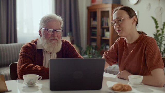 Elderly man with white beard putting on glasses, listening to young daughter explaining how to use Internet on laptop at table in living room