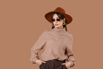 Beautiful young fashionable woman in hat and sunglasses on brown background