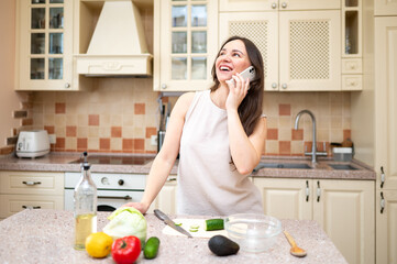 young woman in bright kitchen talking on phone and smiling, planning to cook salad. Fresh vegetables and board are on table