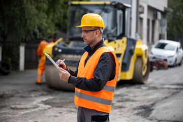 Construction manager oversees road maintenance project while noting details on clipboard in urban...