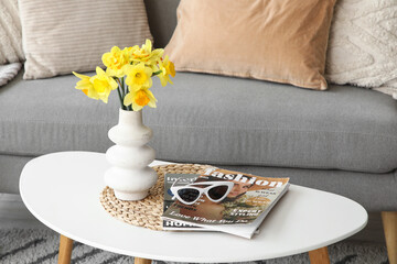 Vase of beautiful daffodil flowers with sunglasses and magazines on coffee table near sofa in...
