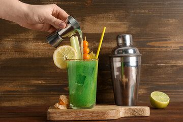 Female hand pouring vodka into glass of green bloody mary cocktail on wooden background