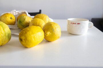 Bright yellow lemons in a white fabric bag on a white indoor table, with a white cup in the background.