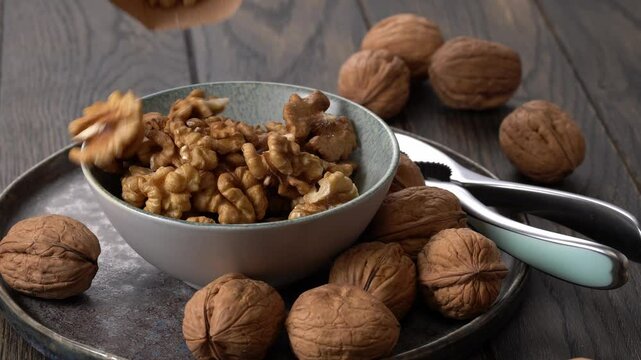 Bowl of walnuts being poured into it from unseen container surrounded by whole walnuts on wooden table.