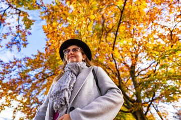 Happy middle aged woman in black brimmed hat walking against autumn tree background