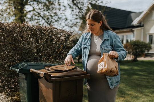 Young pregnant woman throwing garbage in trash can at back yard