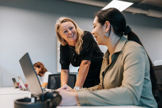 Happy blond businesswoman talking with coworker sitting at desk in office