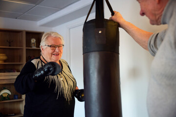 Happy senior woman wearing gloves punching on bag while talking with male friend in rehabilitation center