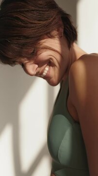 An active adult woman stands by a window with natural light, showcasing a beautiful smile, embodying health and a sporty lifestyle.